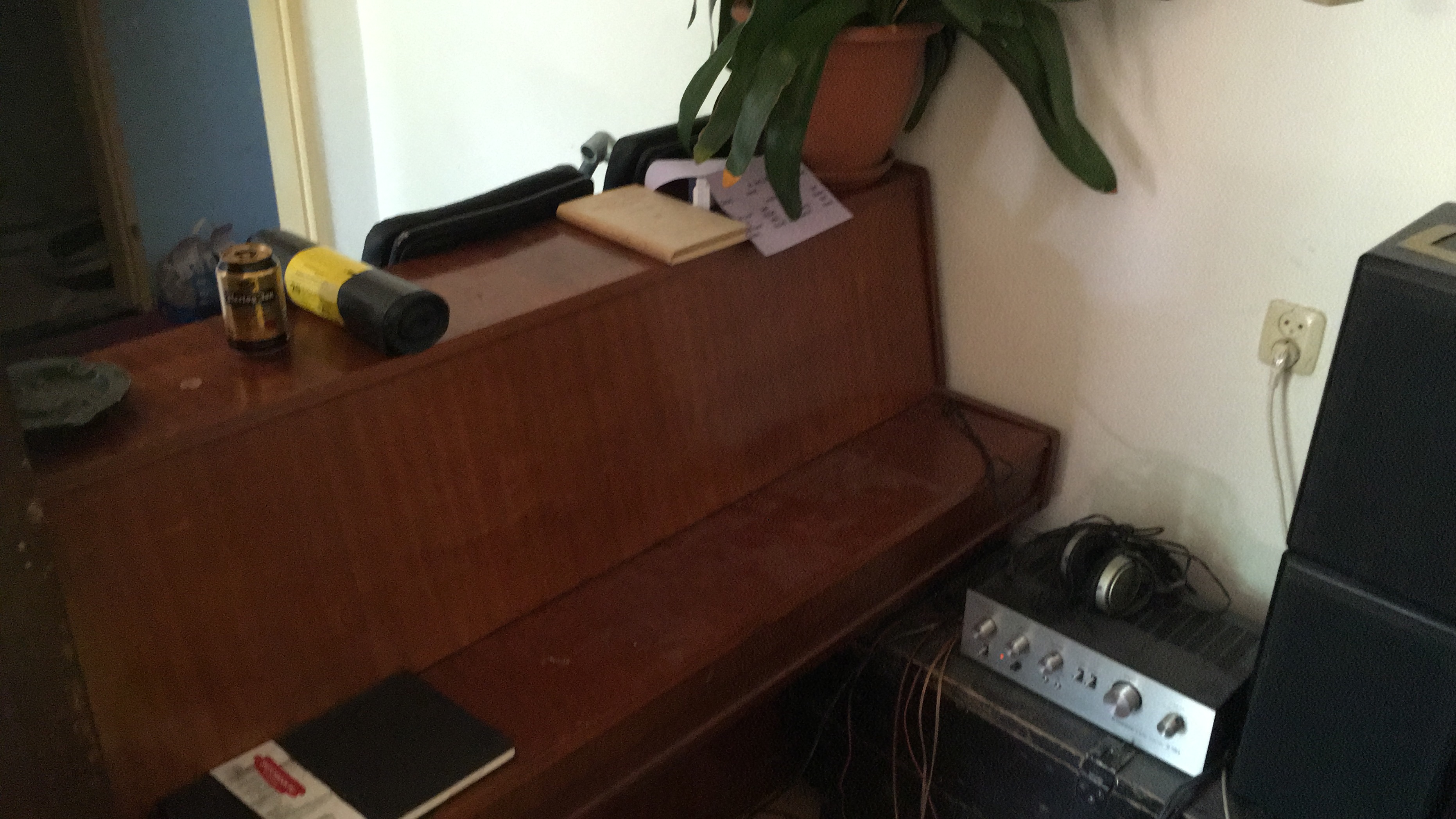 A wooden piano serves as a creative workspace adorned with a can of beer, a rolled-up yellow and black mat, a small notebook with some loose papers, and a potted plant. To the right of the piano, an amplifier with headphones resting on it is visible, along with a power outlet featuring a connected plug.