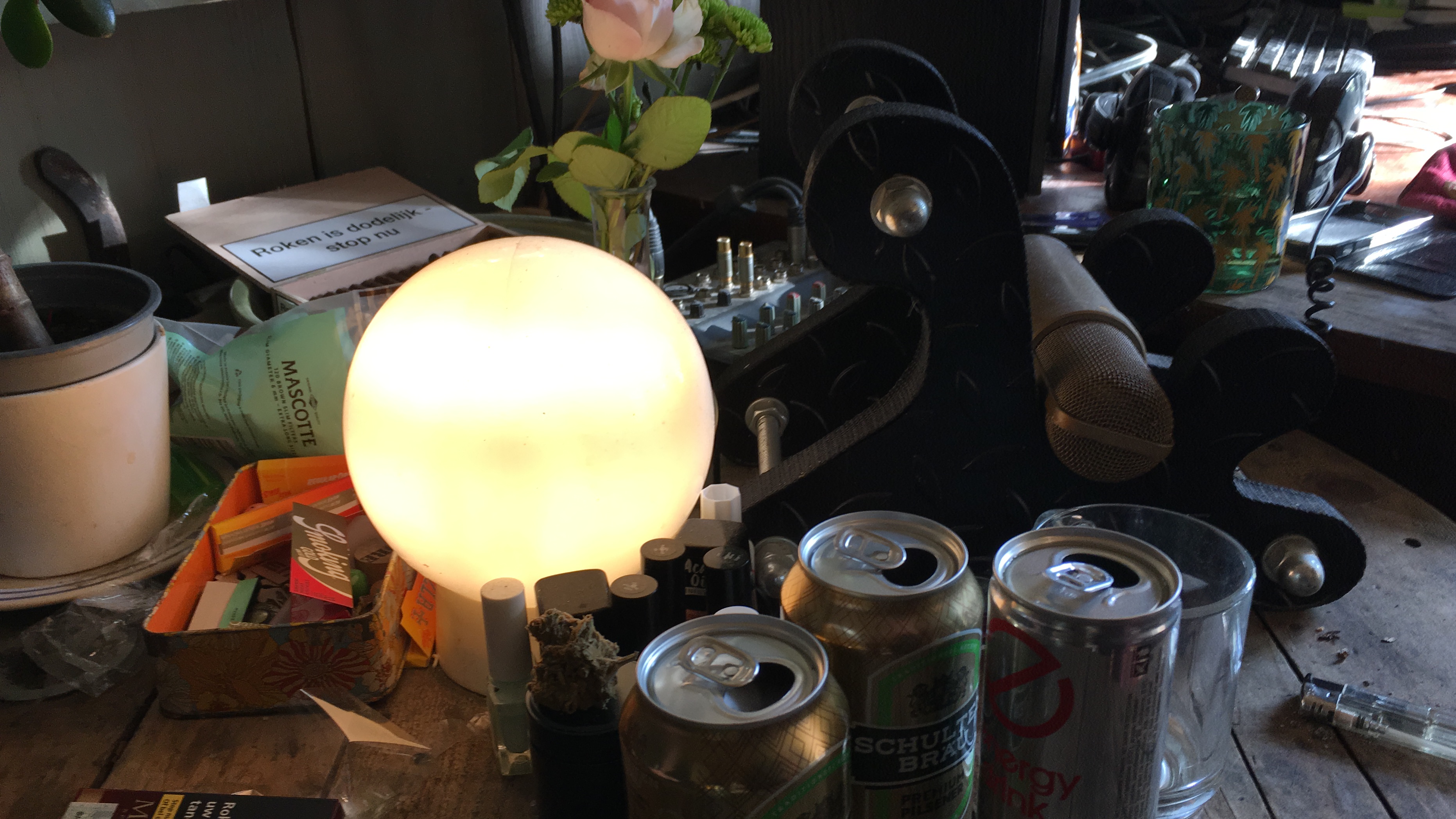 Cluttered tabletop scene centered on a glowing spherical lamp. Surrounding the lamp are diverse objects: a small potted plant on the left; a box with the message 'Roken is dodelijk - stop nu'; a packet labeled 'MASCOTTE'; a tin box holding matchboxes and papers; several cans including one marked 'Schulten Brau' and another 'Energy Drink'; a glass cup; a microphone on a stand; a mixing console with multiple knobs and cables; a green glass featuring a floral pattern; headphones; and various small items like nail polish bottles, a lighter, and a container with what appears to be a cannabis bud. This eclectic arrangement evokes a creative workspace blending musical elements with everyday life.