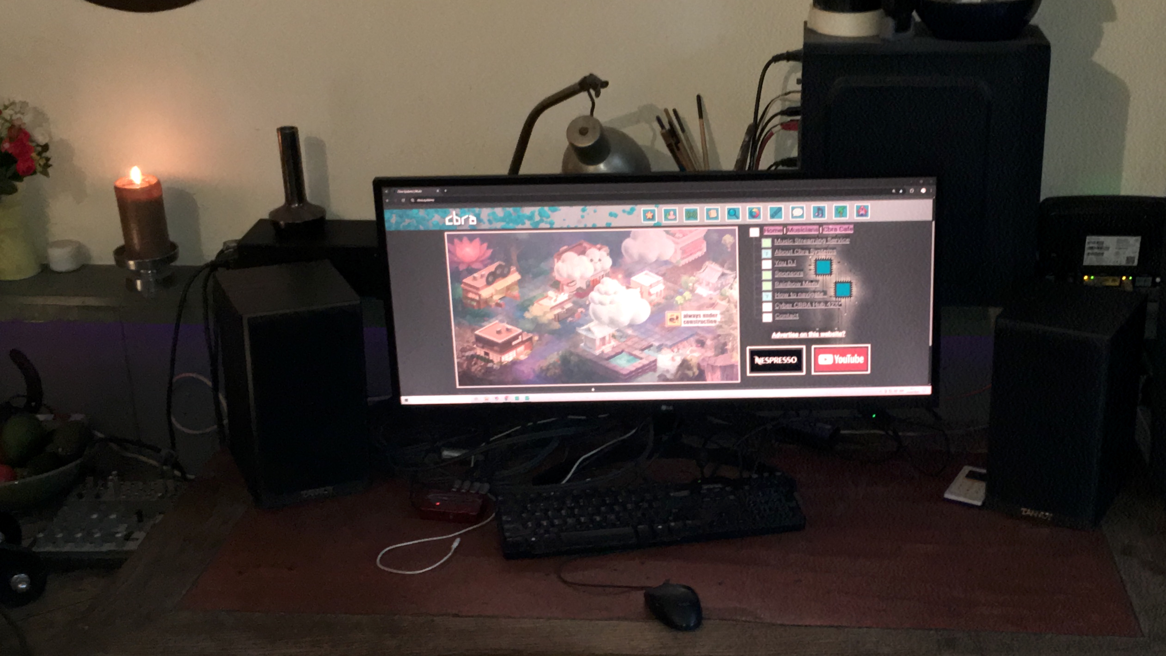 A vibrant home office workspace featuring a computer desk with a widescreen monitor displaying an interactive website. The site shows a colorful, cartoonish map alongside a menu listing options such as Home, Applications, Extra Credit, Music Streaming Service, About CBRA, You DJ, Resources, Banking, Calendar, and Advertise at this website, with logos for Nespresso and YouTube visible. The desk includes a keyboard, mouse, and various cables, while to the left a shelf holds a burning candle, a vase with flowers, and a small white object. On the right, speakers, a lamp, and other electronic devices complete the creative setup.