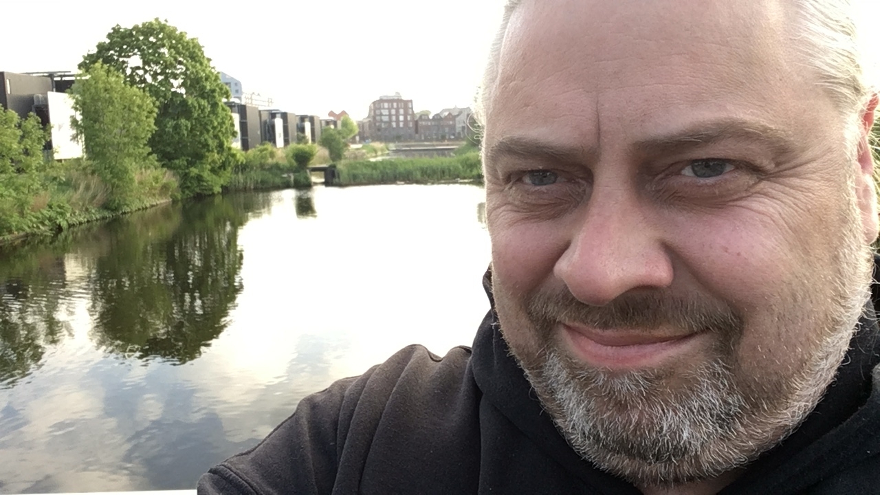 A man smiling in a close-up selfie with a calm canal, green trees, and modern buildings in the background on a clear day.