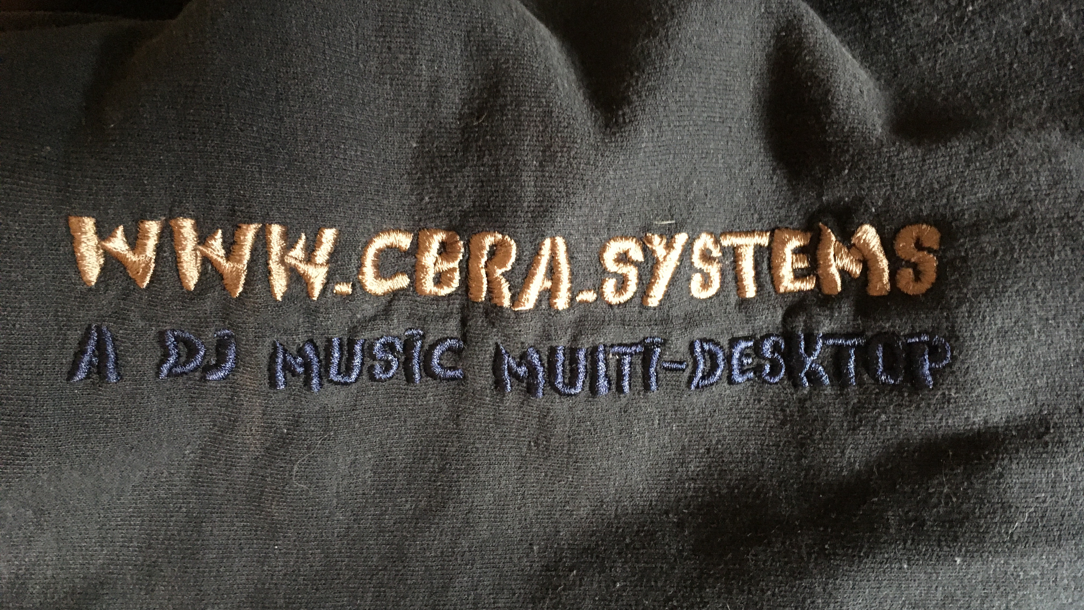 Close-up of black fabric featuring a two-line embroidered design. The top line displays warm tan text that reads 'WWW.CBRA.SYSTEMS', and the bottom line shows dark blue text stating 'A DJ MUSIC MULTI-DESKTOP.' The tactile embroidery stands out against the dark background, merging modern digital branding with handcrafted elegance.
