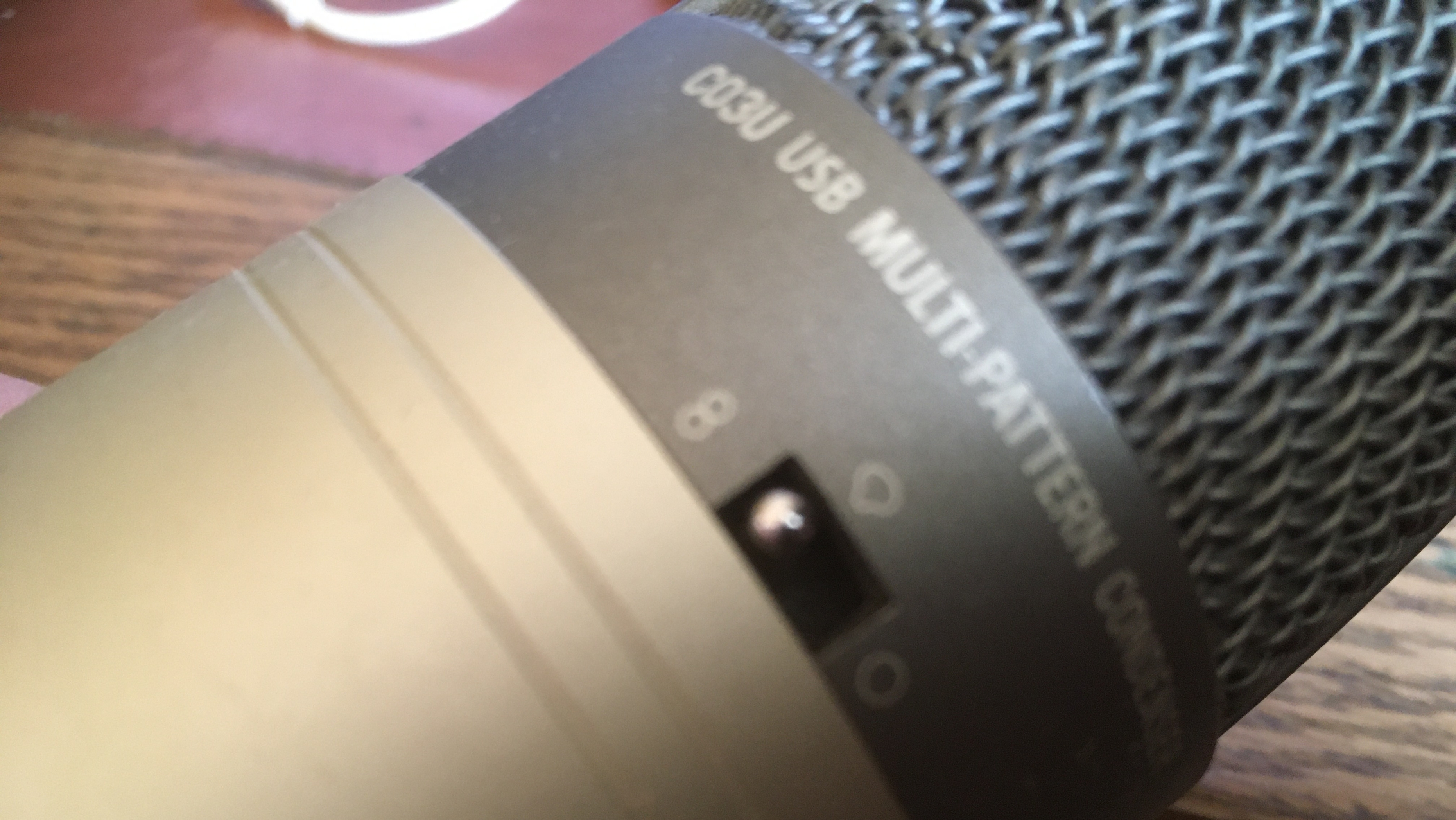 Close-up view of a USB multi-pattern condenser microphone with a metallic mesh grille on a beige and black body. The microphone displays the text 'C03U USB MULTI-PATTERN CONDENSER' and features a three-position switch labeled '2', 'O', and an infinity symbol, highlighting its versatility in sound capture.
